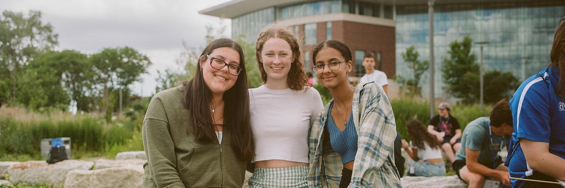 Three students smile at camera on campus lawn in front of university hall.